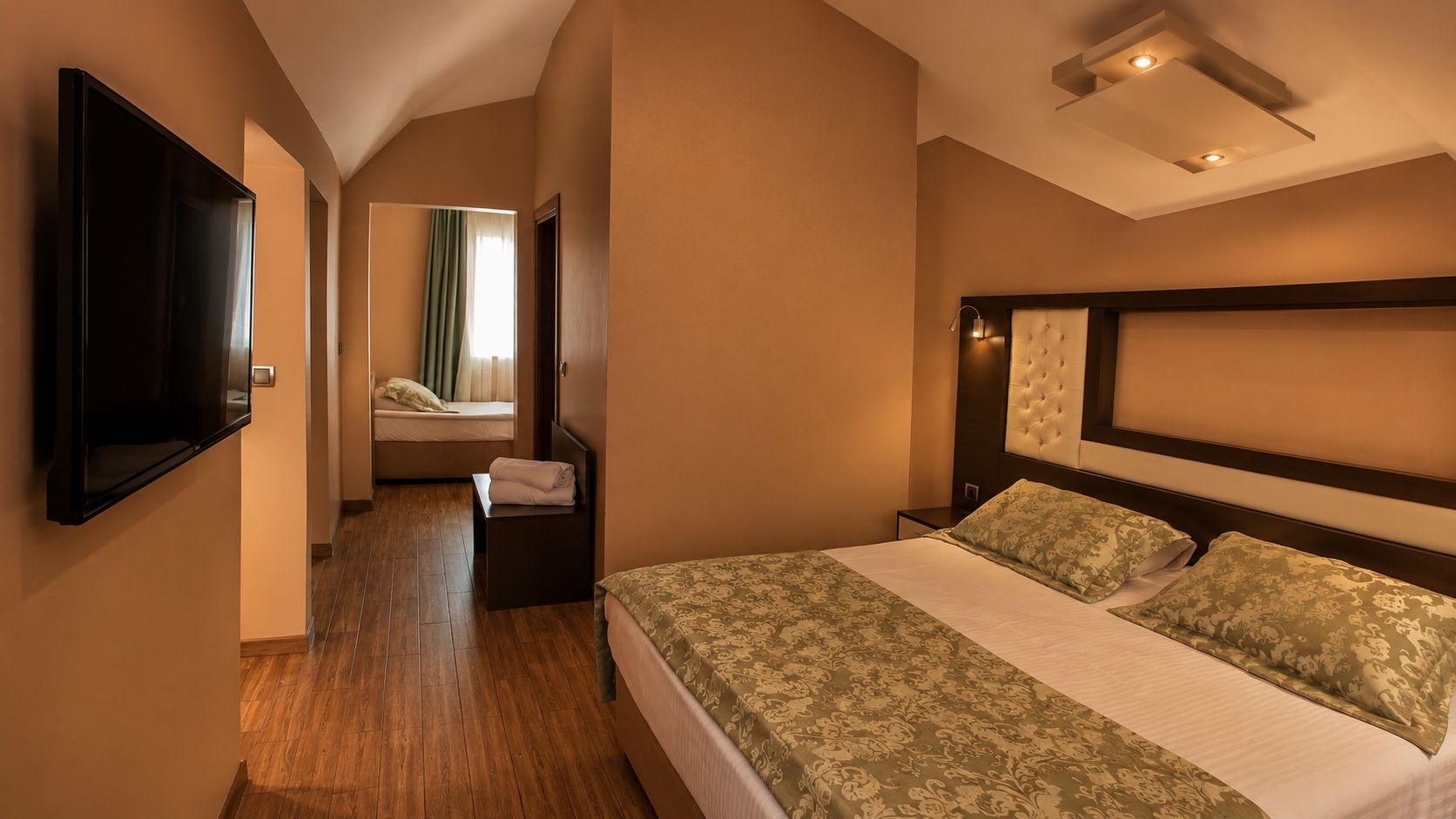 A hotel room with a large bed, a flat-screen TV mounted on the wall, a bedside table, and a window with curtains.