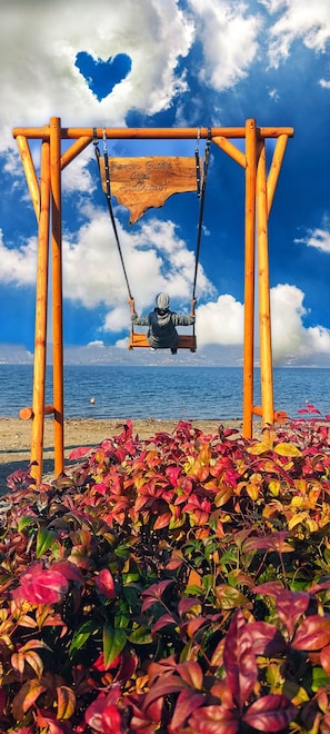 Un portique en bois avec une découpe en forme de cœur dans le ciel, une plage avec des plantes aux couleurs vives au premier plan, et un ciel bleu clair parsemé de nuages.