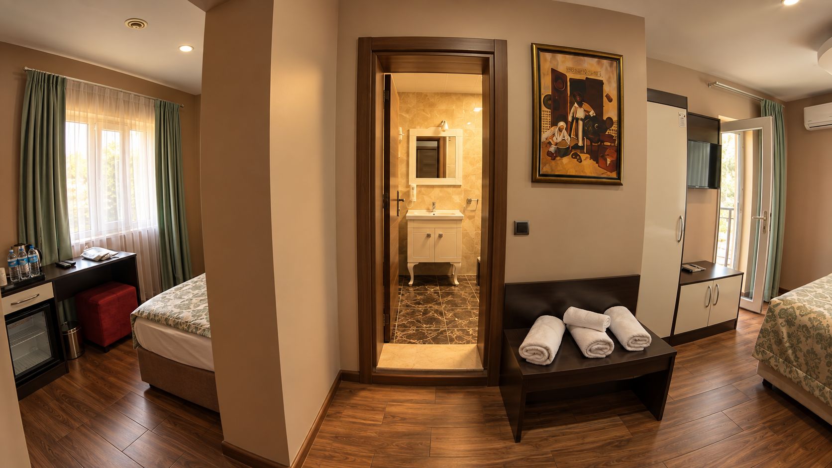 A hotel room with a bed, a desk, a chair, a small table with towels, a mirror, and a painting on the wall.