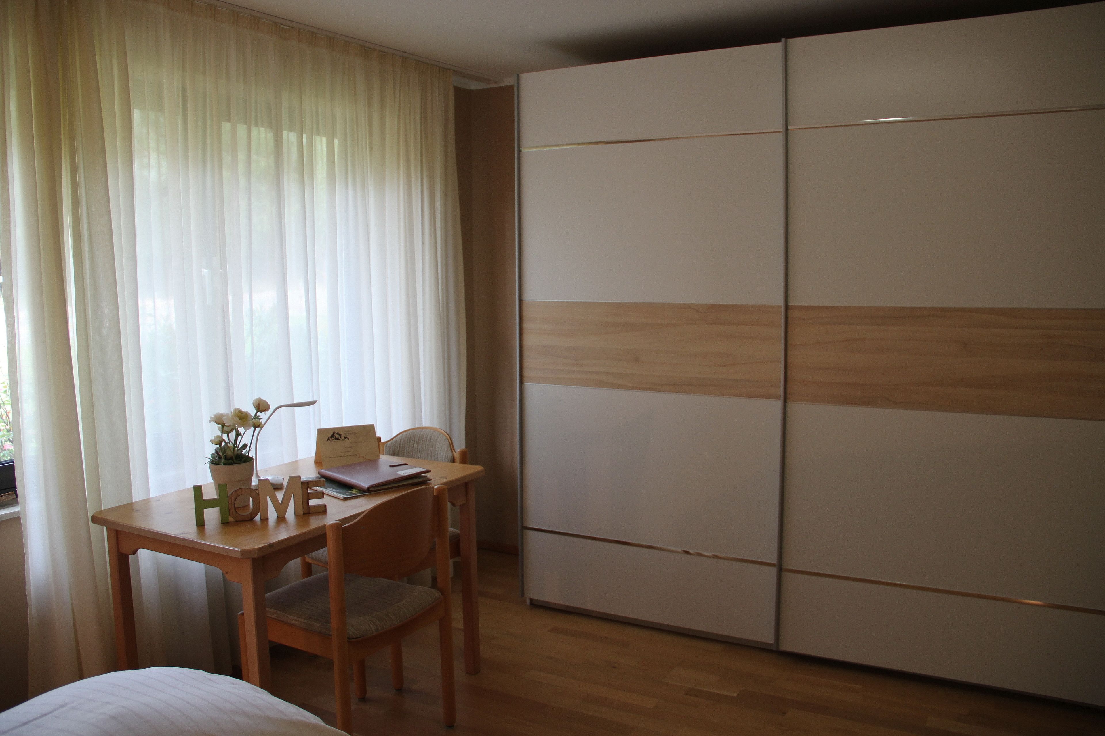 A bedroom with a wooden desk, chair, and a bed. There is a wardrobe with sliding doors and a window with sheer curtains.
