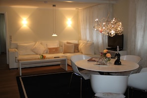 A modern living room with a white sofa, a coffee table, and a round dining table with chairs. There is a chandelier above the dining table and a flat-screen TV mounted on the wall. - Verenas Parkhotel (Löffingen)