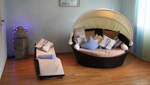 Una habitación con piso de madera, un sillón redondo de mimbre con dosel, un sofá con cojines, una mesita con un adorno, y un cuadro colgado en la pared.