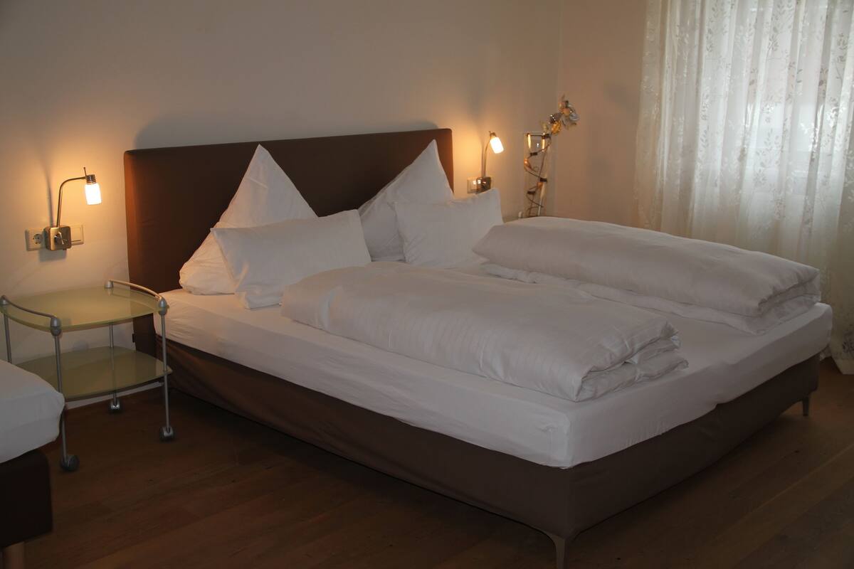 Un lit bien fait, avec du linge de lit blanc, une table de chevet avec une lampe et un luminaire fixé au mur.