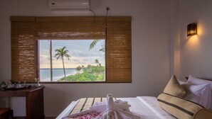 A hotel room with a bed, a desk, and a window with a view of palm trees and the ocean. - Ananya Beach Resort (Rekawa)