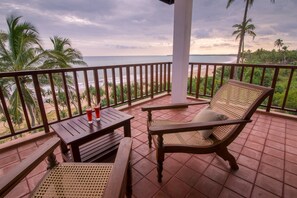 A wooden deck with a table and chairs, overlooking a beach with palm trees and the ocean.