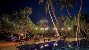 An outdoor dining area with tables and chairs, surrounded by palm trees and a pool. - Ananya Beach Resort (Rekawa)