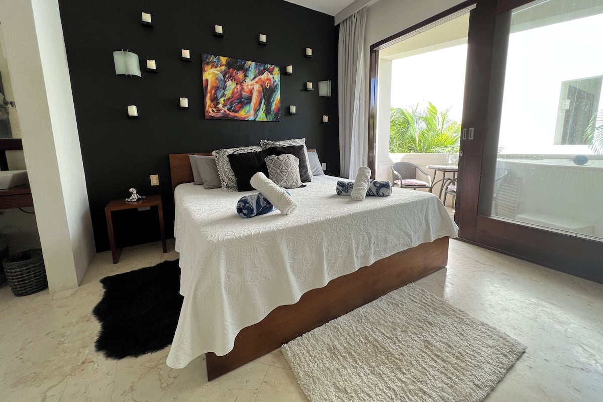 A modern bedroom with a large bed, a painting on the wall, and a view of greenery outside.