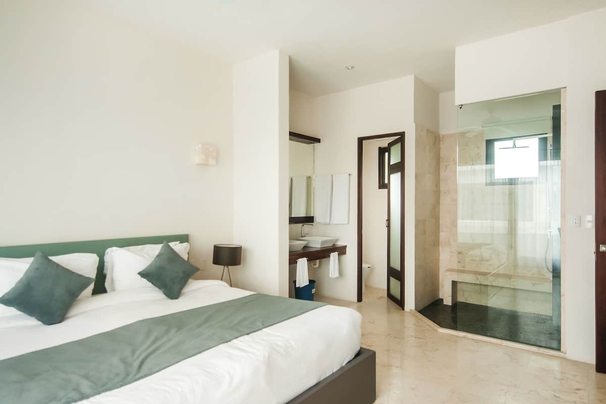 A modern hotel room with a large bed, a bathroom with a glass shower enclosure, and a visible sink area.