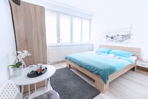 A modern bedroom with a wooden bed, a small bedside table, a white chair, a round table with a tray, and a window with sheer curtains. - The Dots Hostel (Zagreb)