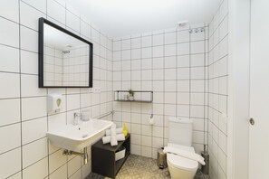 A bathroom with white tiled walls, a white sink with a mirror above it, a toilet, and a small shelf with toiletries. - BYPILLOW Wander (Barcelona)