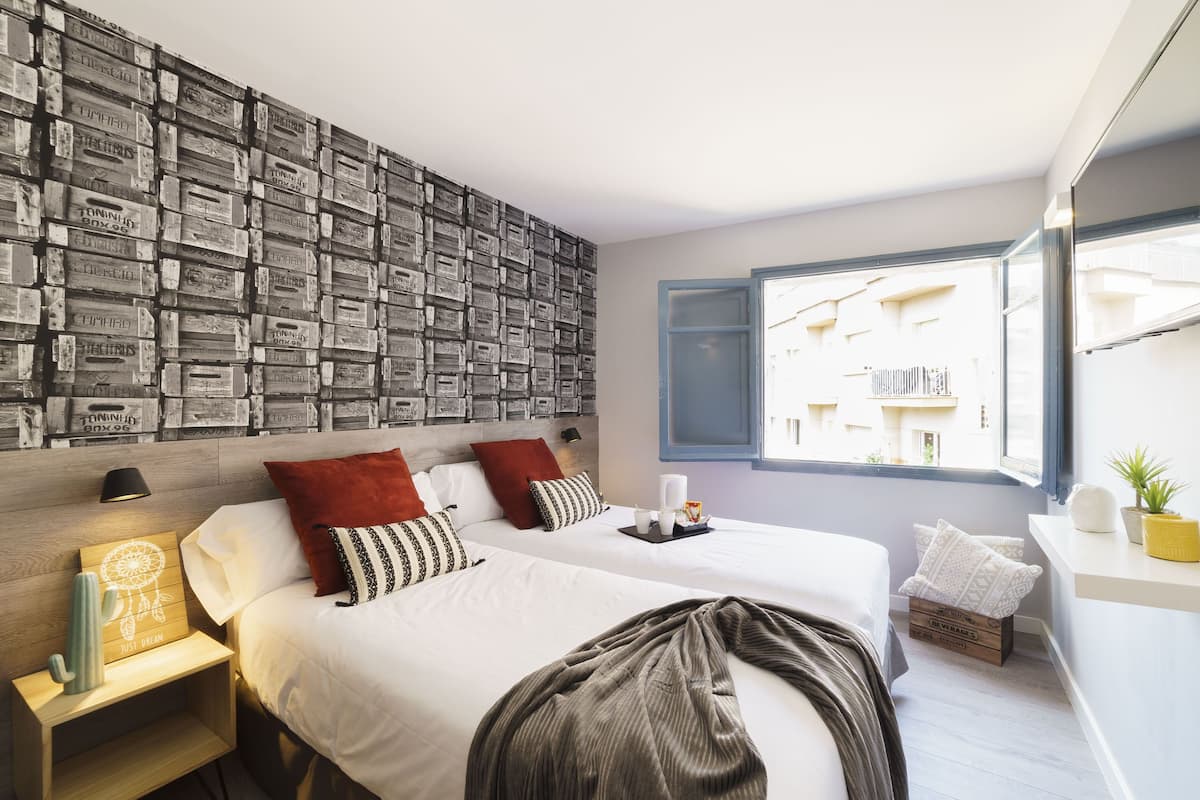 A hotel room with two beds, a wall of stacked crates, a window with a view, and a bedside table with a lamp and a decorative item.