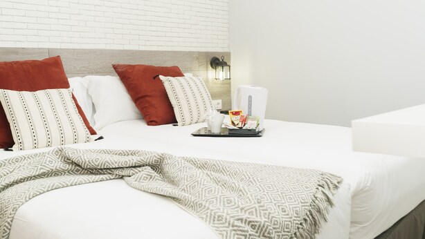 A hotel room with a white brick wall, a bed with white bedding and patterned pillows, a bedside table with a tray of breakfast items, and a wall-mounted lamp. - BYPILLOW Wander (Barcelona)