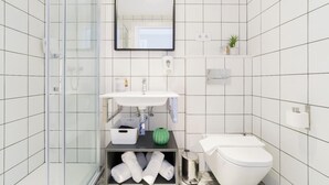 A modern bathroom with a glass shower enclosure, a white sink, a toilet, and a small shelf with toiletries. - BYPILLOW Wander (Barcelona)