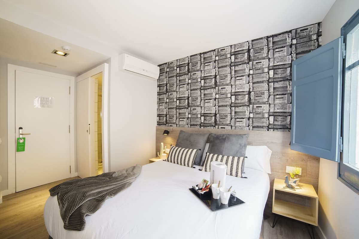 A hotel room with a neatly made bed, a bedside table with a lamp, a wall decorated with vintage-style tiles, and a window with blue shutters.
