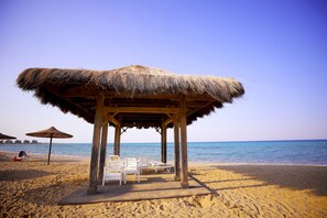 A beach hut with a thatched roof, white lounge chairs, and a wooden deck.