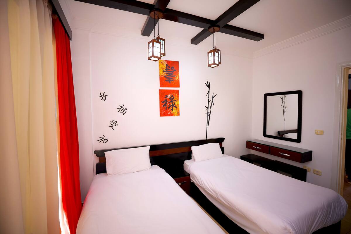 Une chambre d’hôtel avec deux lits, un rideau rouge, un ventilateur de plafond et des décorations murales comprenant de la calligraphie et un tableau encadré.