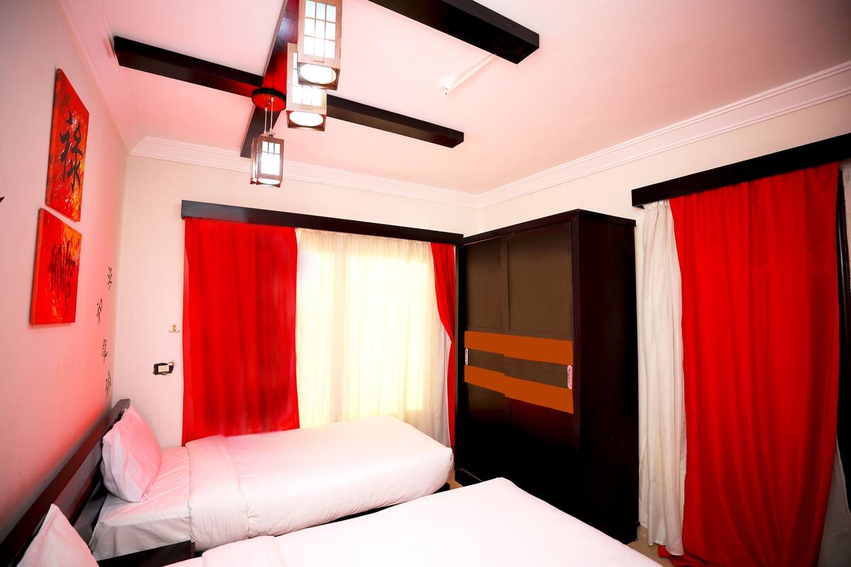 Une chambre d’hôtel avec deux lits, des rideaux rouges, un ventilateur de plafond et des œuvres d’art accrochées aux murs.