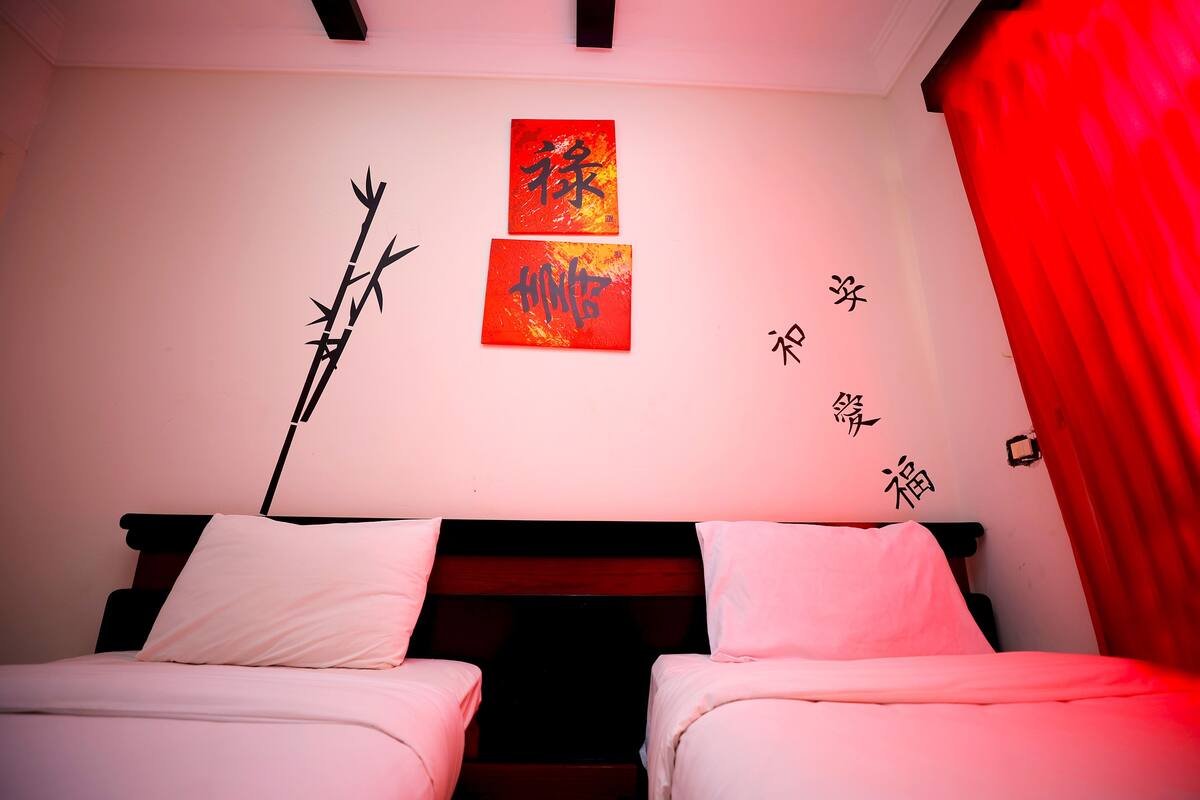 Une chambre avec deux lits, un rideau rouge et des œuvres d’art murales ornées de caractères chinois.