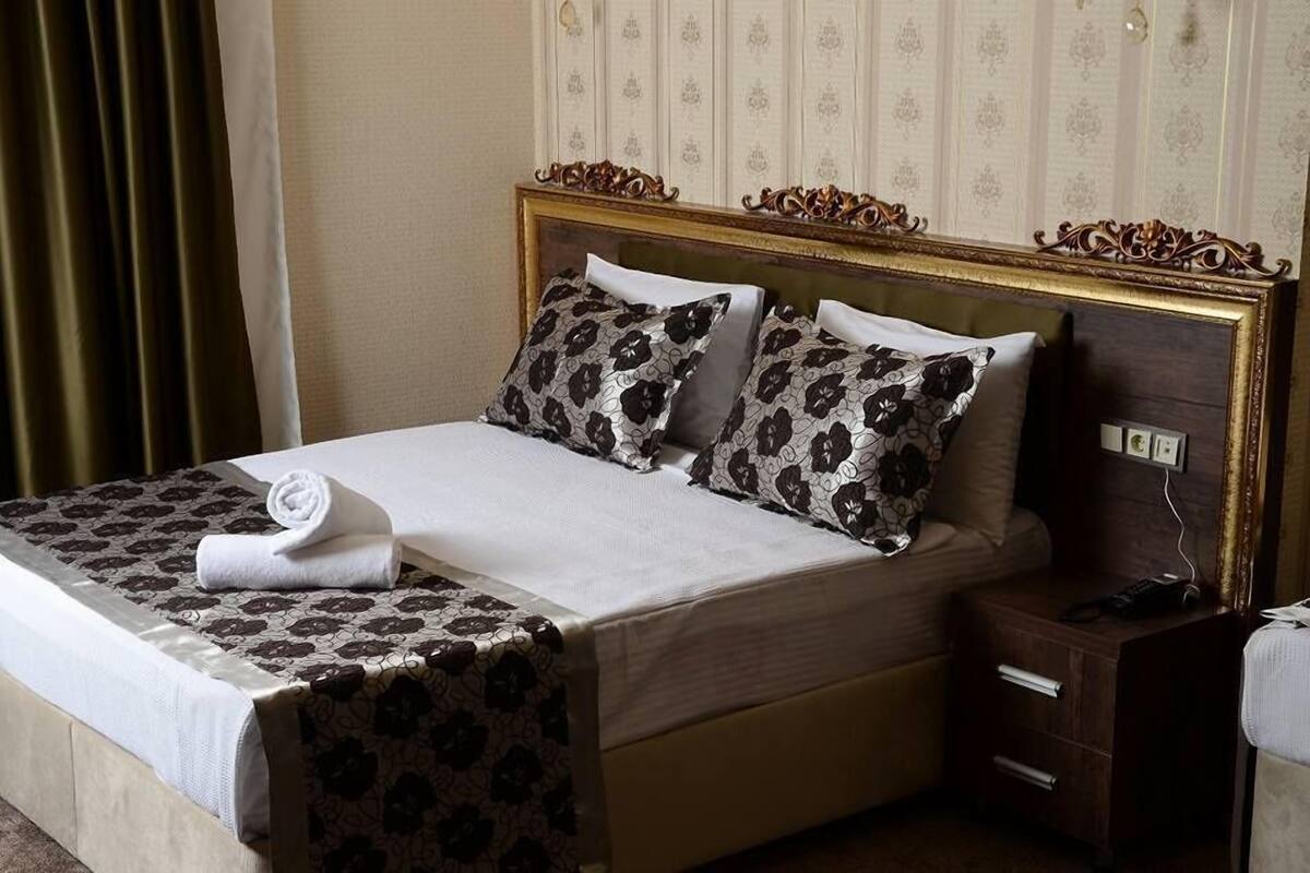 Una cama bien hecha con ropa de cama estampada, una mesita de noche de madera con un teléfono y una pared con un panel decorativo.