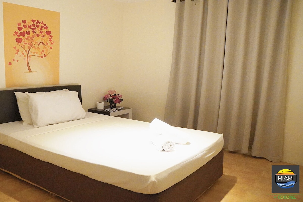 A neatly made bed with white linens, a dark headboard, and a wall art of a tree with hearts.