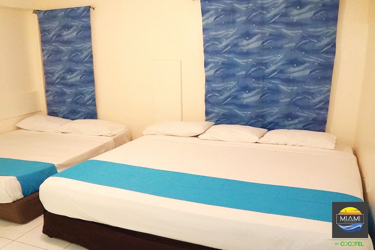 Two single beds with blue and white bedding, a blue curtain, and a Miami-themed logo on the wall.