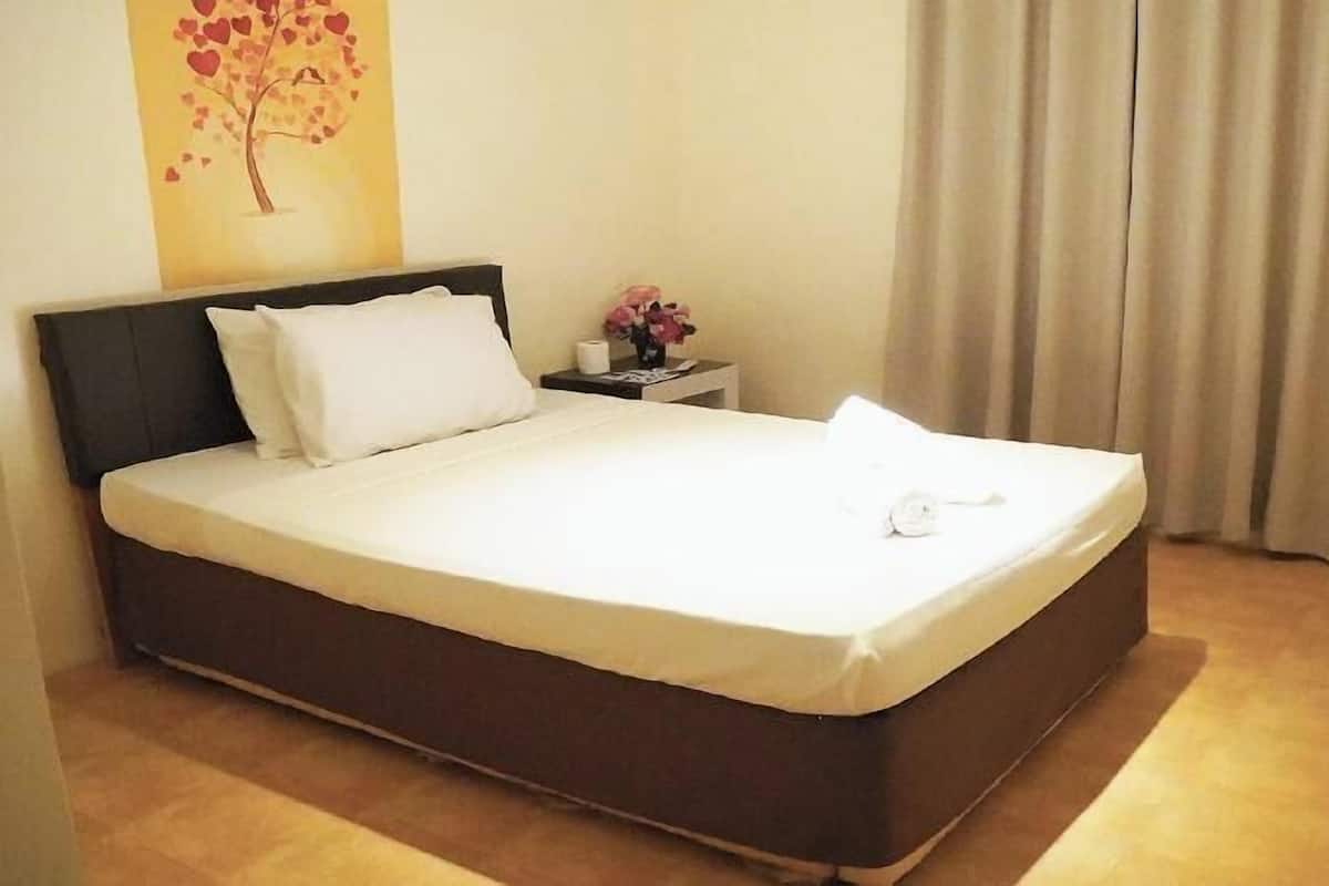A neatly made bed with white linens, a dark headboard, a small bedside table with a vase of flowers, and a wall painting of a tree with hearts.