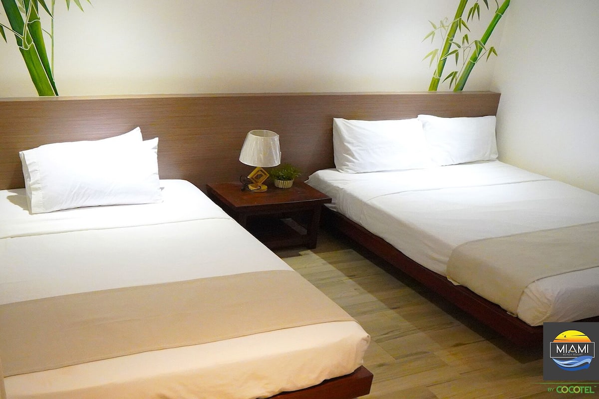 A hotel room with two single beds, a nightstand with a lamp, and a small plant.