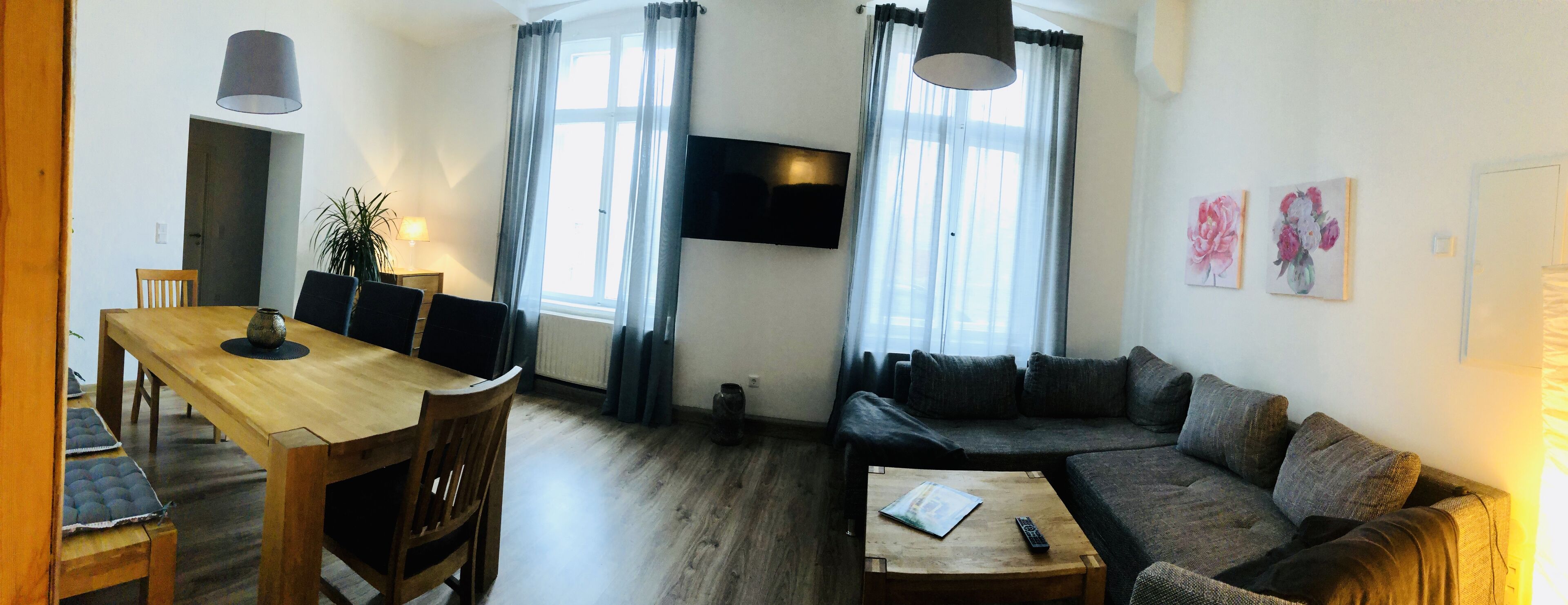A living room with a wooden dining table, a sofa, and a TV mounted on the wall.