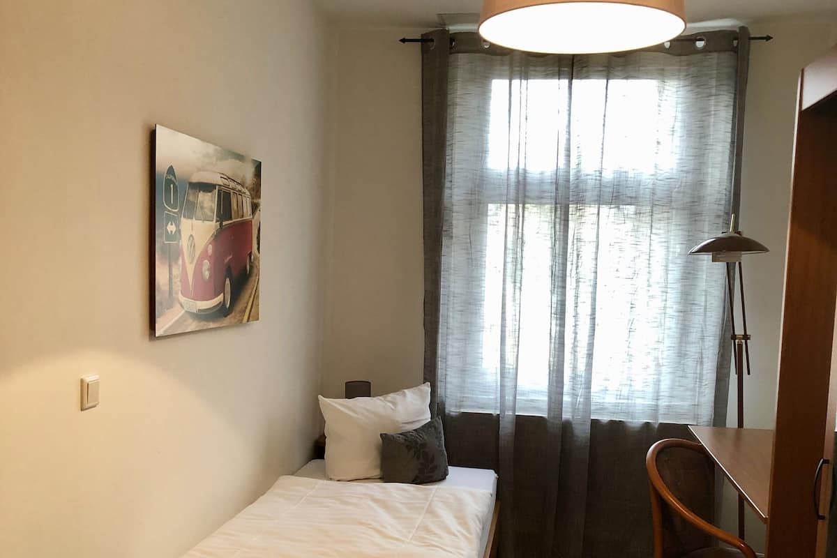 A small, neatly arranged bedroom with a single bed, a chair, a desk, and a painting on the wall.