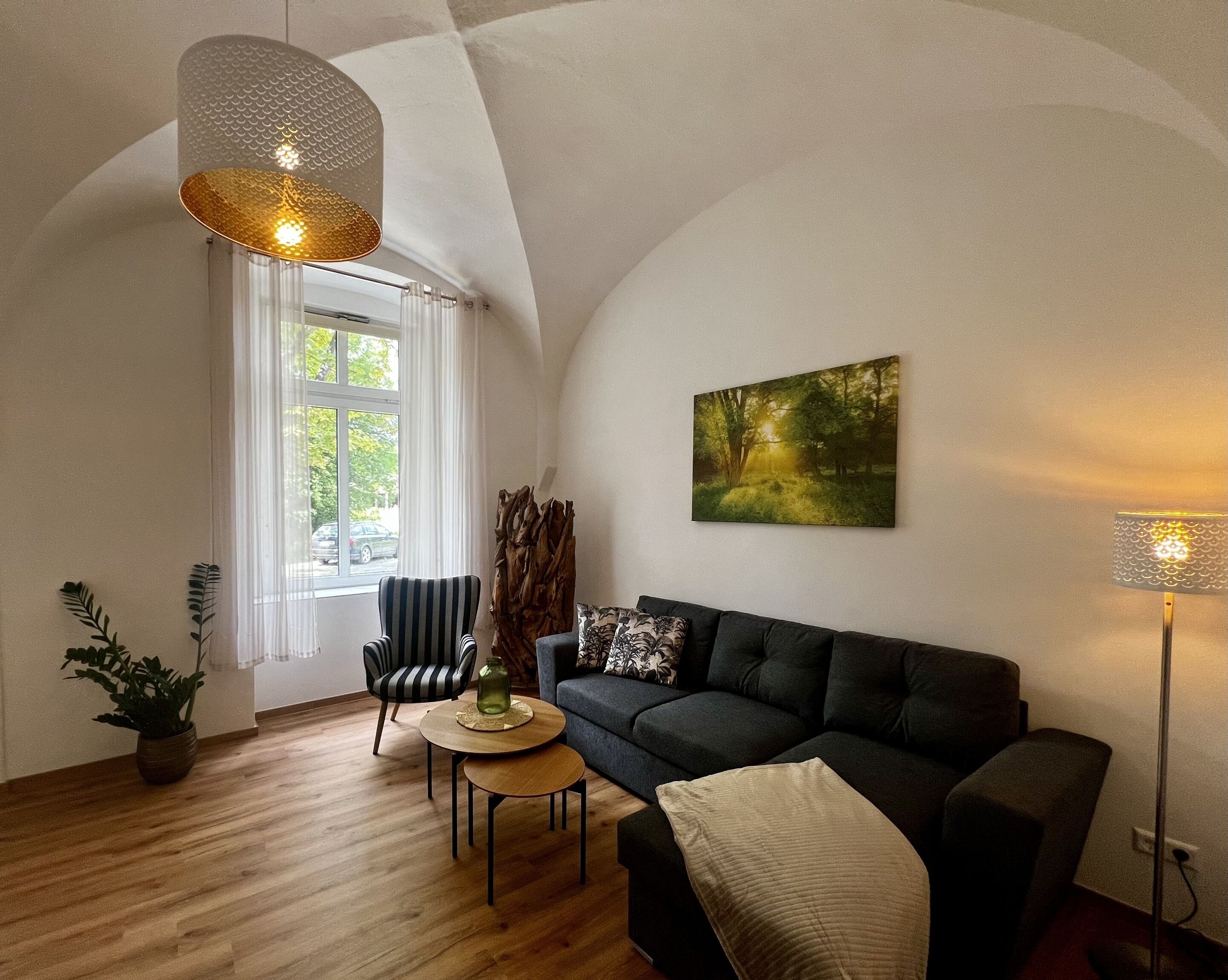 Una sala de estar acogedora con un sofá oscuro, una mesa redonda de madera, un sillón a rayas y un ventanal con cortinas.