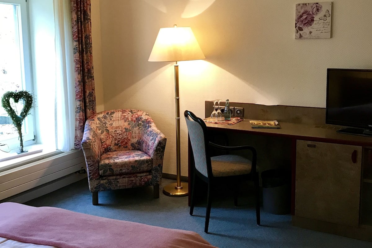 A hotel room with a bed, a desk with a chair, a television, a lamp, and a window with curtains.