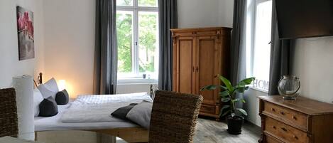 A bedroom with a bed, a dresser, a television, a window with curtains, and a plant.