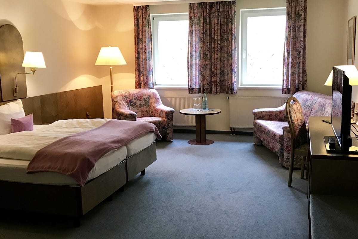A hotel room with a large bed, two armchairs, a small round table, and a desk with a lamp.