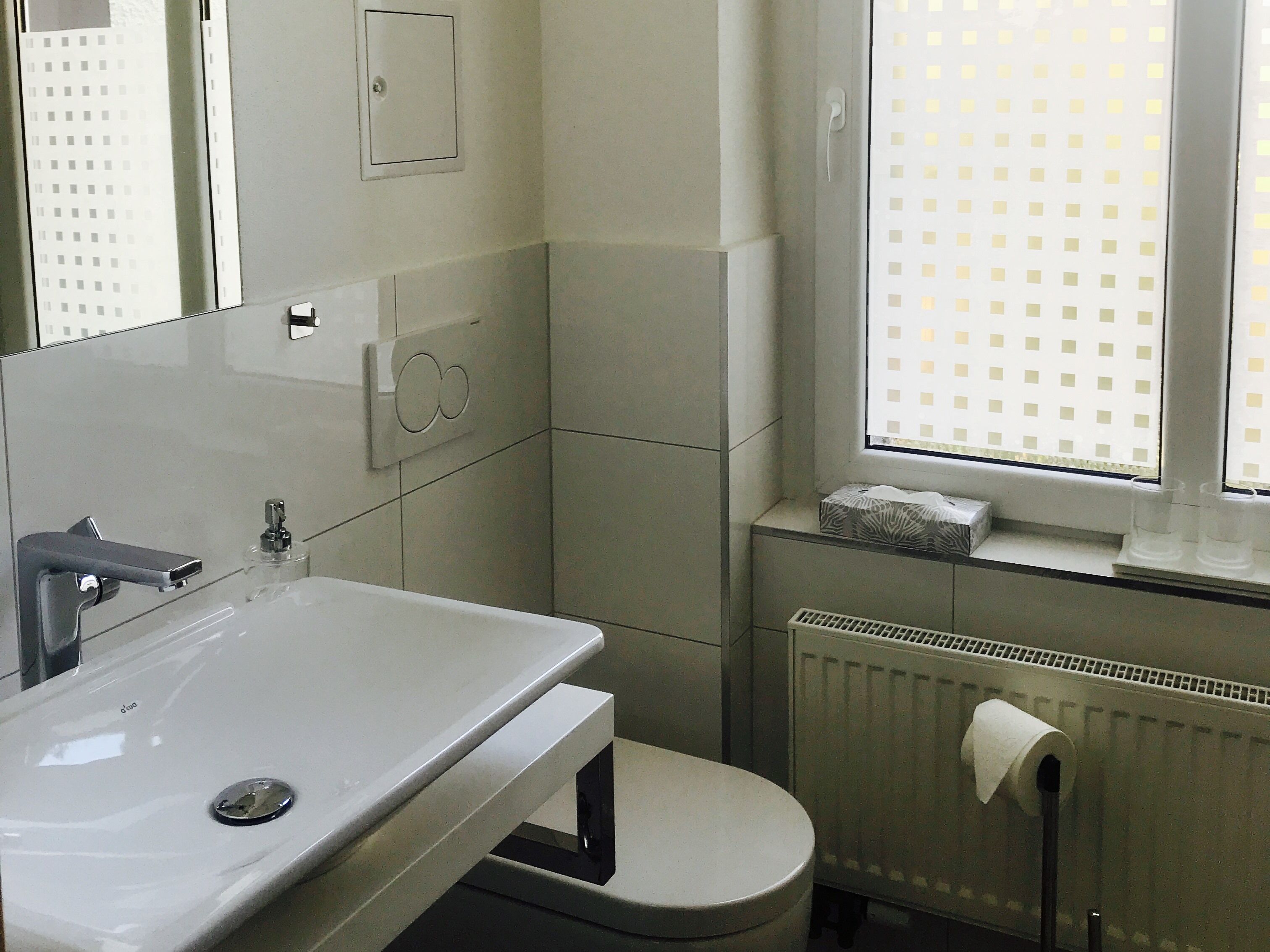 Un baño con un lavabo blanco, un inodoro, un radiador y una ventana con diseño de cuadrícula.