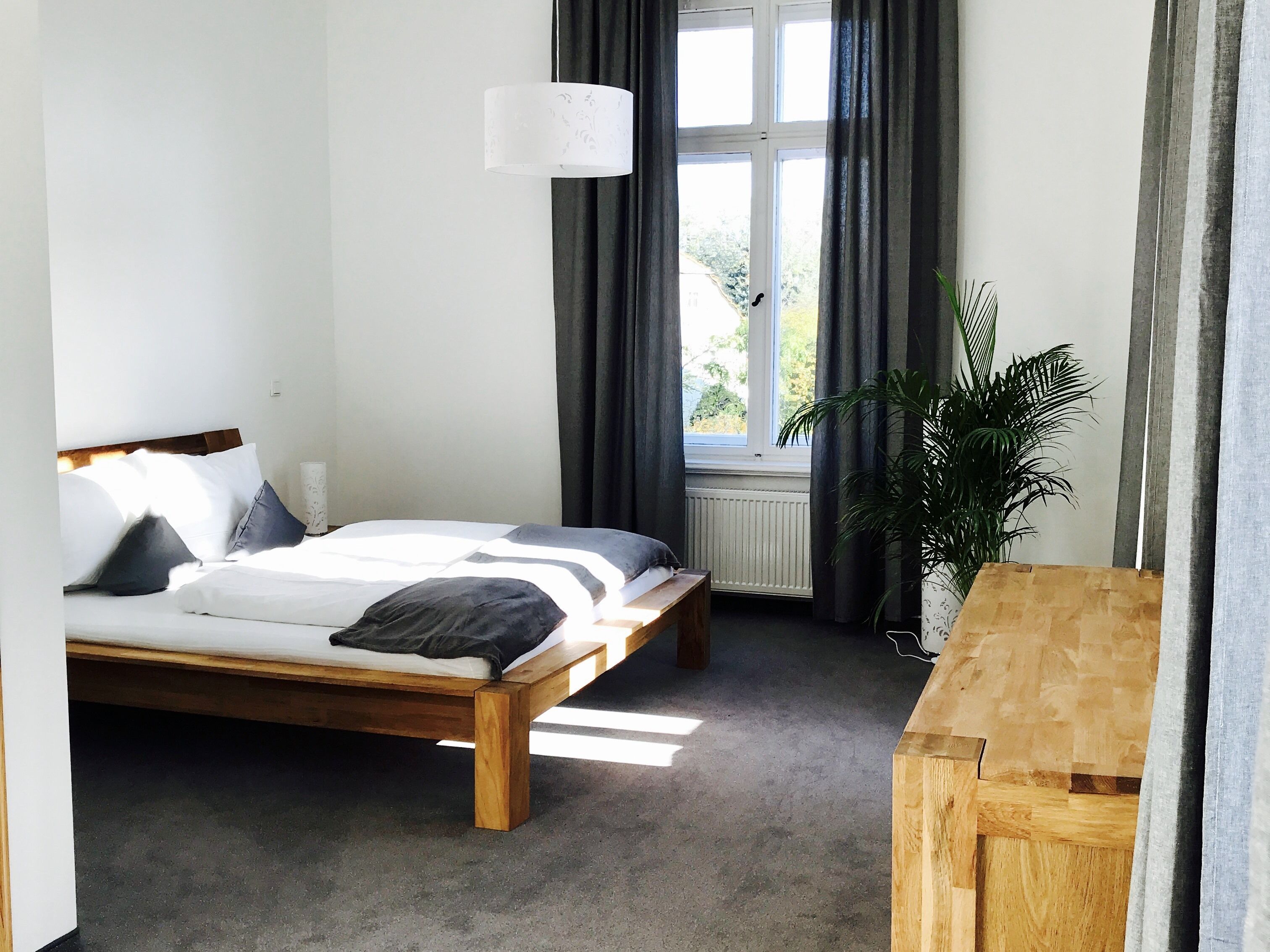 A bedroom with a bed, a bedside table, a lamp, a window with curtains, and a potted plant.