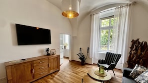 A spacious living room with a wooden floor, a flat-screen TV mounted on the wall, a wooden cabinet, a striped armchair, a round coffee table, and a large window with curtains. - Hotel Dein Gutshof (Görlitz)