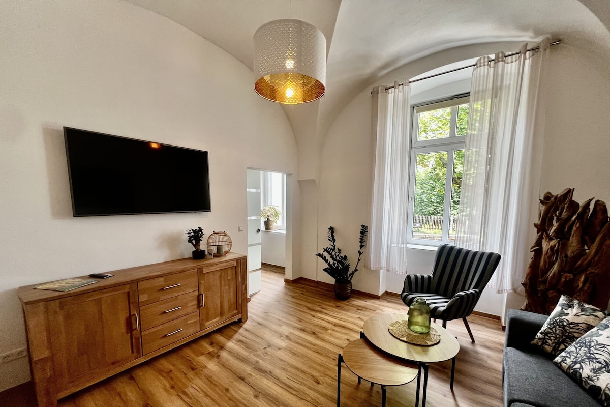 A spacious living room with a wooden floor, a flat-screen TV mounted on the wall, a wooden cabinet, a striped armchair, a round coffee table, and a large window with curtains.