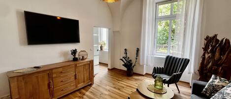 Amplia sala de estar con piso de madera, televisor de pantalla plana montado en la pared, un mueble de madera, un sillón a rayas, una mesa de centro redonda y un gran ventanal con cortinas.