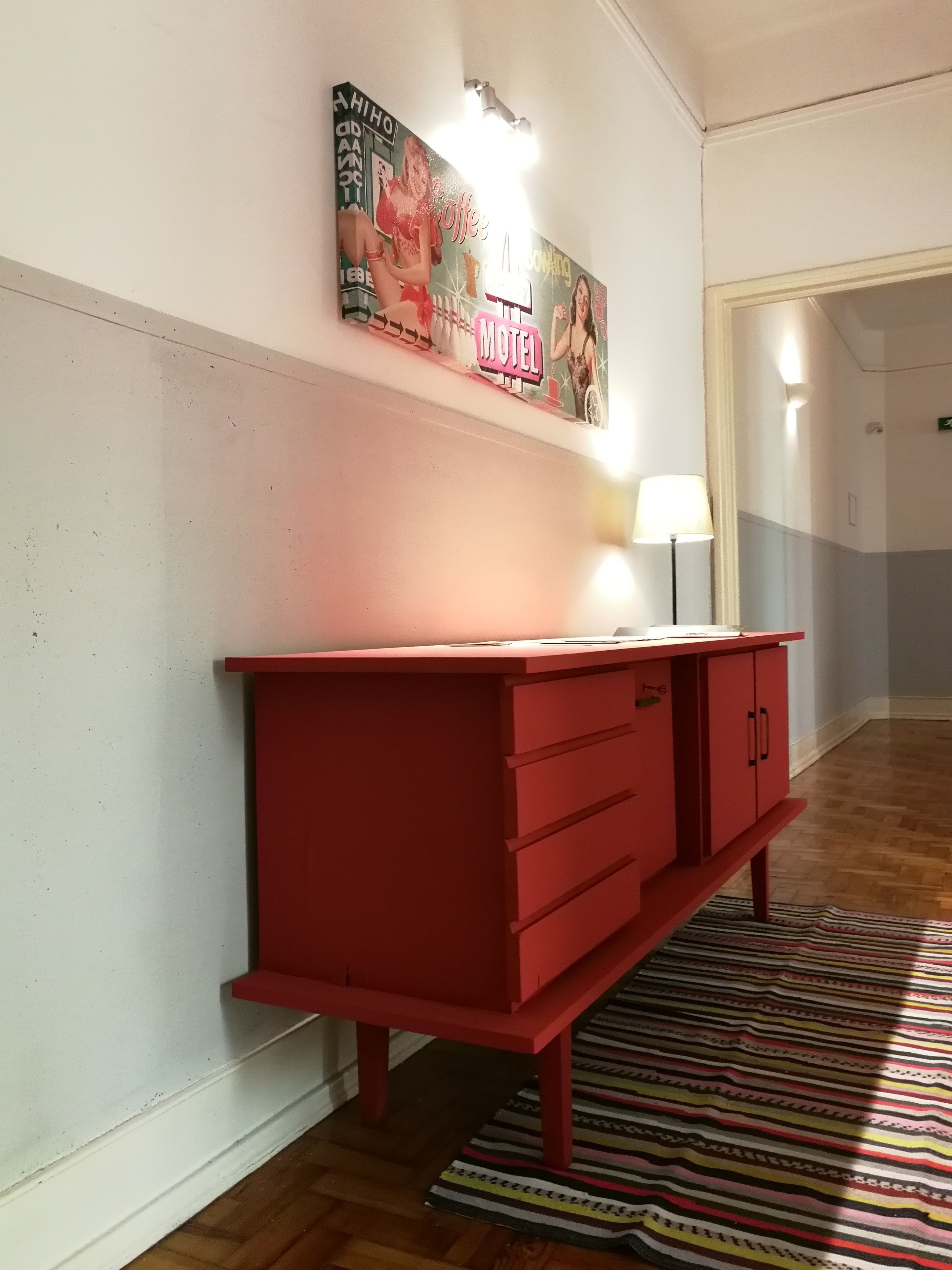 A red dresser with drawers and a mirror, a striped rug on the floor, and a wall with a neon sign and a picture.