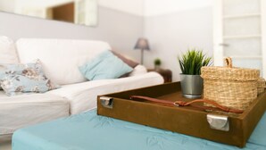 Una habitación con un sofá blanco, una pequeña planta en maceta, una cesta de mimbre y una maleta marrón sobre un edredón azul.