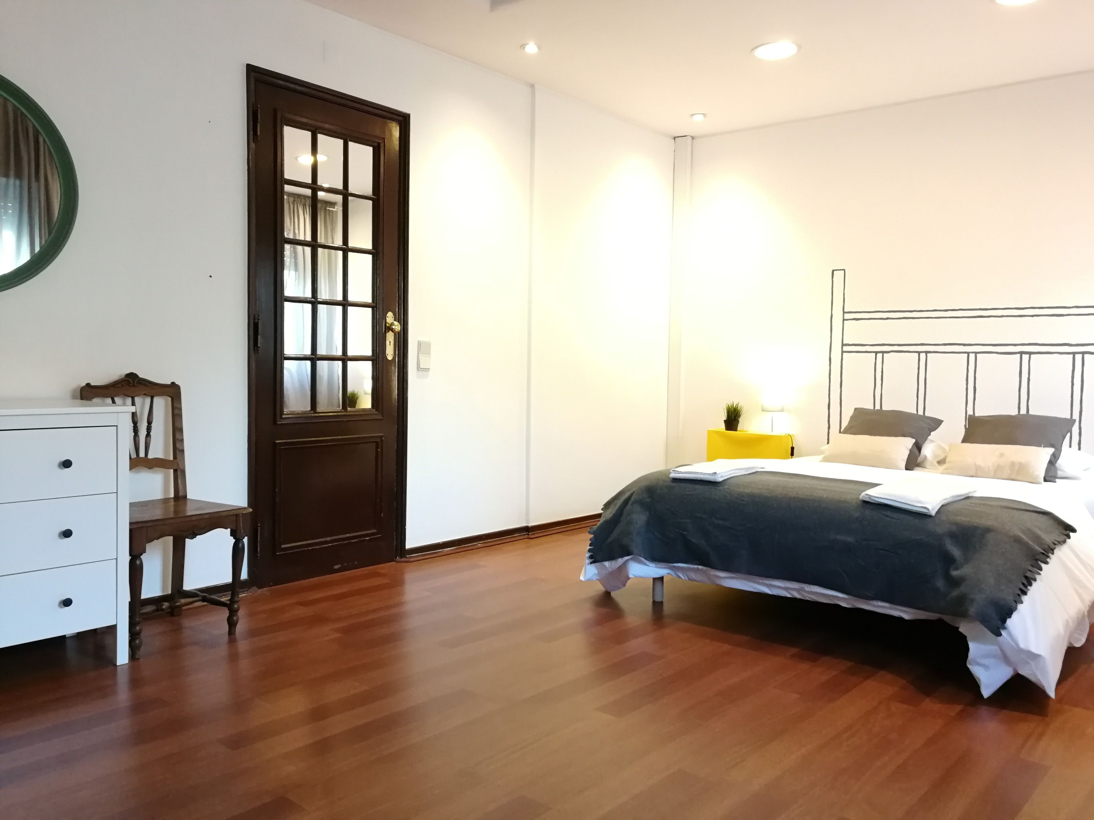 Une chambre avec un plancher en bois, un lit recouvert d’une couverture foncée, une table de chevet avec une lampe, une chaise et une commode.