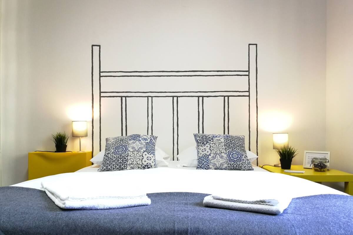 Una cama bien hecha con almohadones estampados, una mesita de noche con lámpara y una pequeña planta sobre una mesita amarilla.