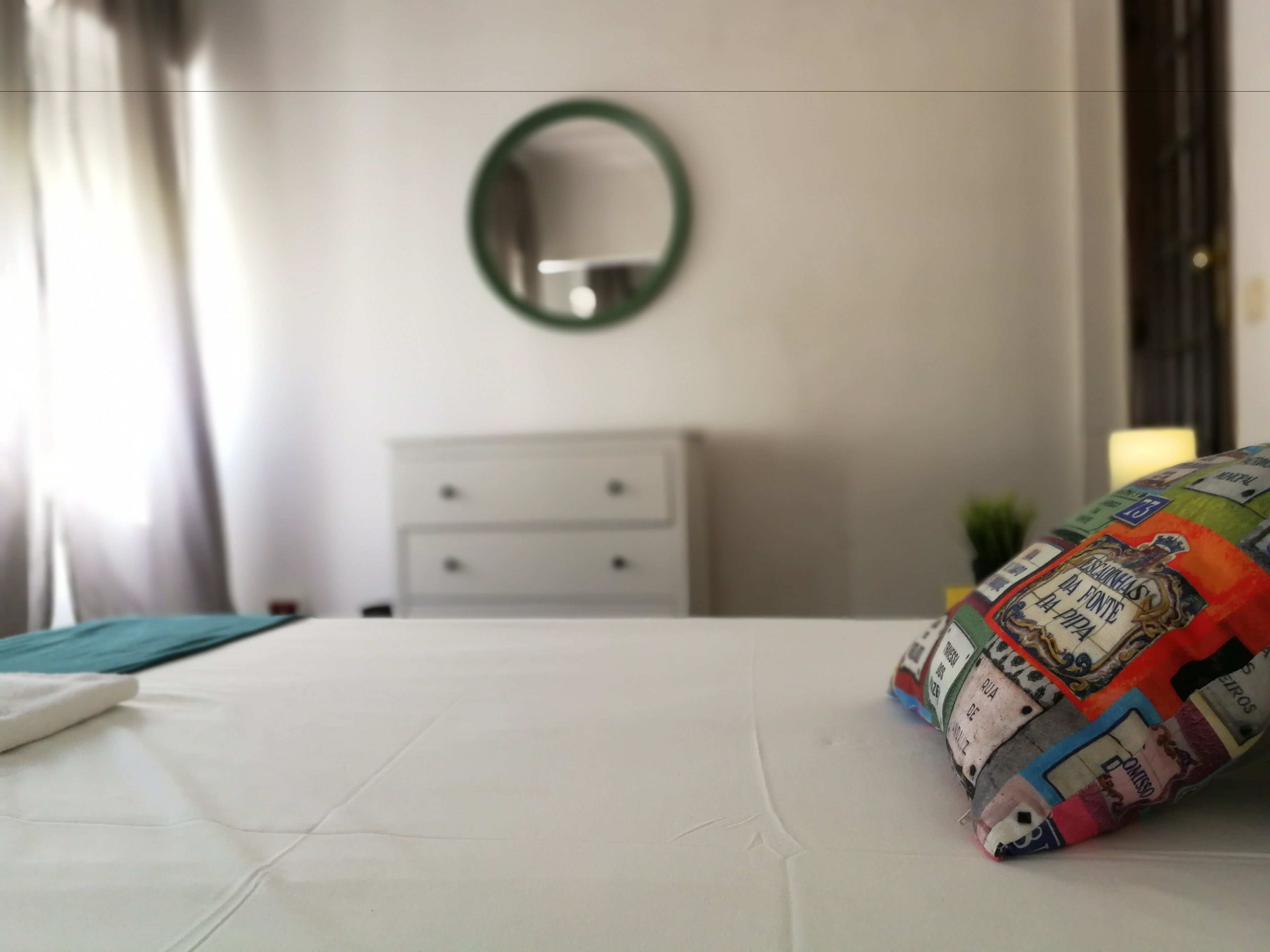 A neatly made bed with a colorful pillow, a white dresser, and a mirror on the wall.