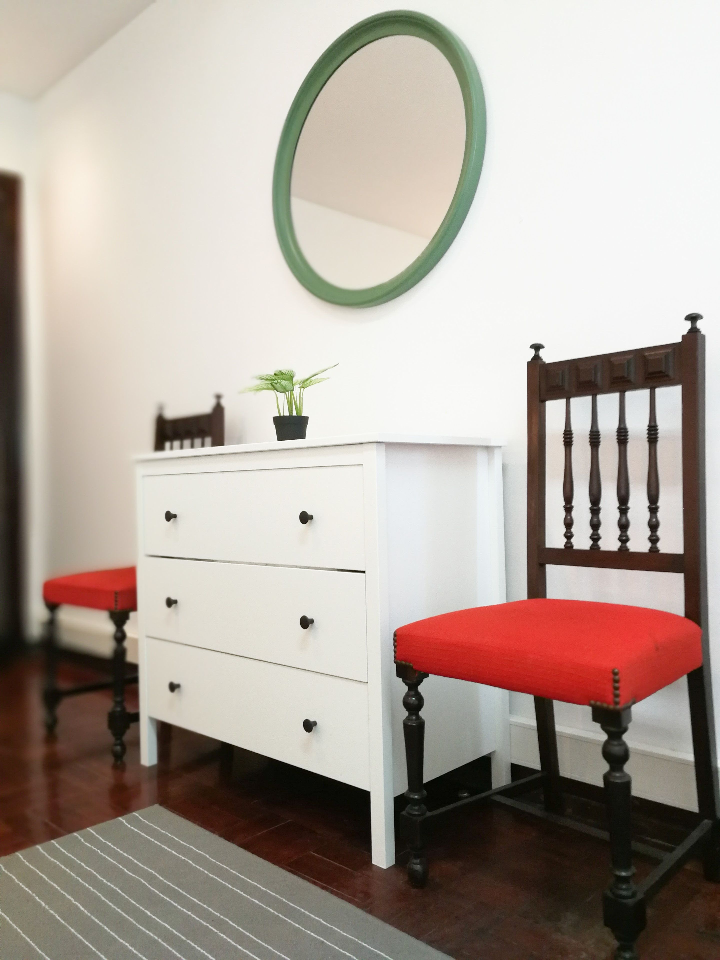 A room with a white dresser, a red chair, a green-framed mirror, and a small potted plant on the dresser.
