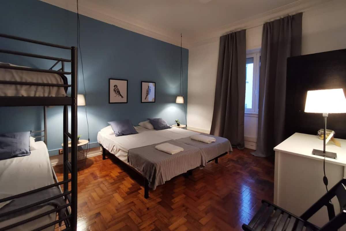 Una habitación con literas, un escritorio, una silla, una cama con almohadas, una lámpara y dos cuadros en la pared.