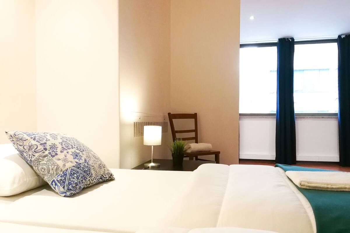 Una habitación de hotel con una cama, una silla, una lámpara y un ventanal con cortinas.