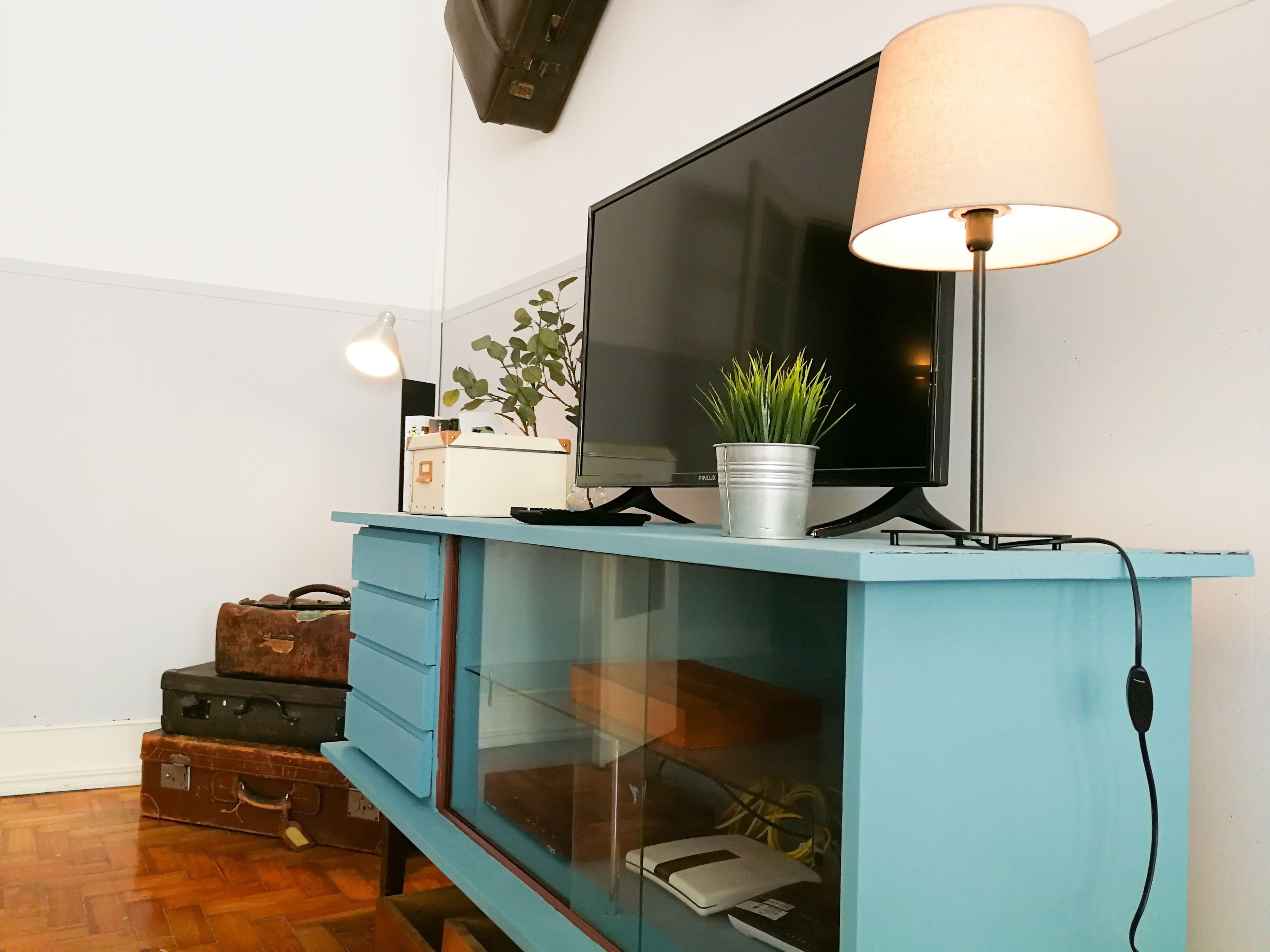 Quarto com televisão em suporte, luminária, planta e armário de vidro com um laptop e mala.