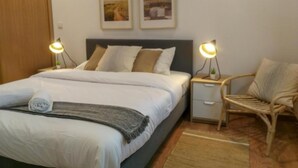 Una cama bien hecha con almohadas y una colcha, una mesita de noche con una lámpara, una silla y dos cuadros en la pared.