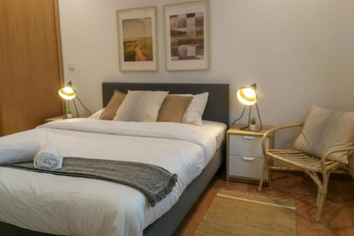 Una cama bien hecha con almohadas y una colcha, una mesita de noche con una lámpara, una silla y dos cuadros en la pared.