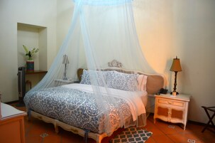 Un dormitorio con cama bajo dosel, mesita de noche con lámpara y una planta pequeña en un estante.